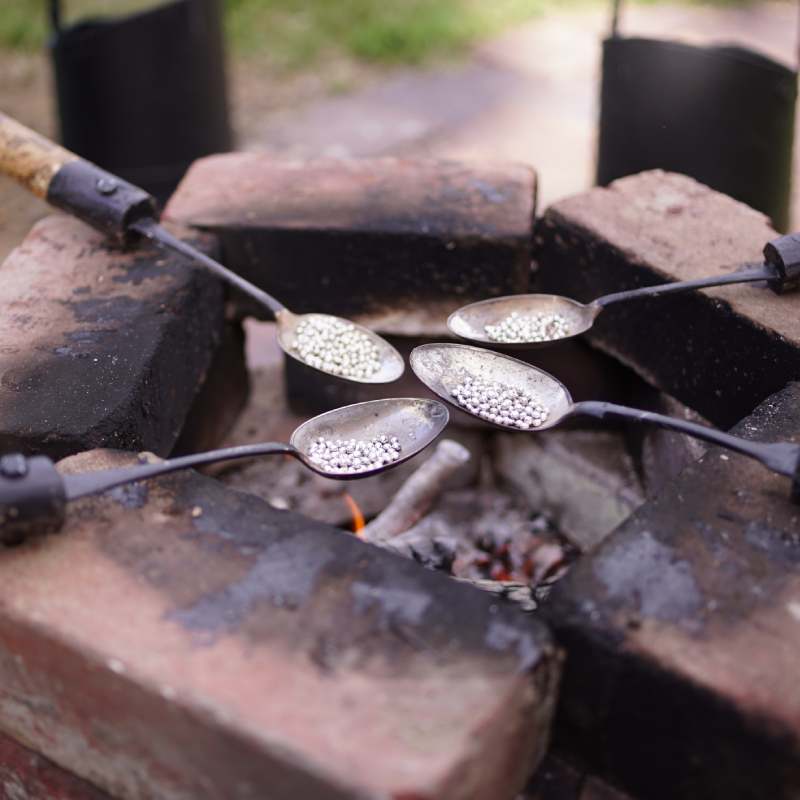 Spoons attached to wooden andles, resting on a square stack of bricks, and fire within, melting pewter silver balls in the cup of each spoon, likely to be poured into molds